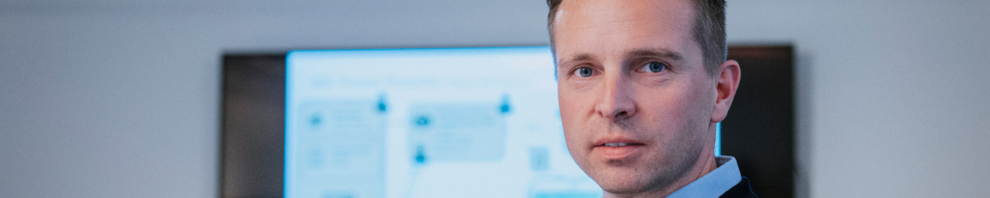 Ein Mann steht vor einem Bildschirm und schaut in die Kamera 