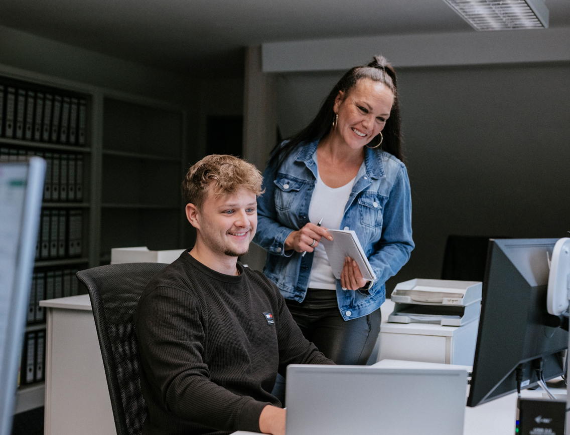 Ein junger Mann sitzt an einem Schreibtisch und eine Frau steht neben ihm.